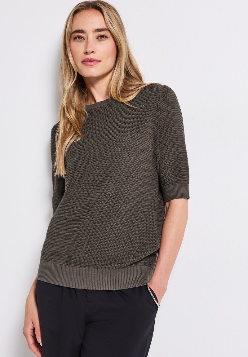 Dark green knitted sweater with short sleeves, textured weave, and ribbed hem. Paired with black pants. Plain background.
