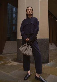 Woman in dark navy suit holding gray drawstring handbag, standing on wet stone pavement near beige and granite building columns.