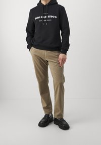 Zwarte hoodie met witte tekst, beige corduroy broek en zwarte schoenen. De hoodie heeft een trekkoord en geribbelde boorden; de broek heeft een verticale textuur.