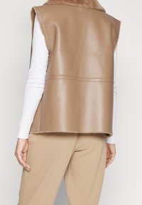 Brązowa kamizelka z ekoskóry z miękkim, puszystym kołnierzem, bez rękawów i surowym wykończeniem. W zestawie z białą bluzką z długim rękawem i beżowymi spodniami.