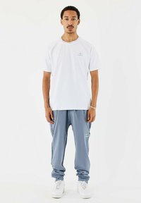 T-shirt bianca a maniche corte con colletto rotondo e logo, abbinata a pantaloni jogger blu con tasche laterali e polsini elasticizzati. Sneakers bianche.