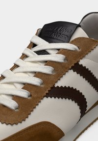 Λευκό δερμάτινο αθλητικό παπούτσι με καφέ σουέτ λεπτομέρειες, ζιγκ ζαγκ διακοσμήσεις και λευκά κορδόνια. Μαύρο λογότυπο στην γλώσσα του παπουτσιού προσθέτει αντίθεση.