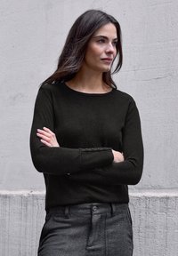 Pull en maille noire avec un col rond et des manches longues, agrémenté de boutons sur les poignets. Porté avec un pantalon gris cintré.