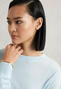 Helle blaue Strickjacke aus weichem Material mit rundem Ausschnitt; sie hat gerippte Bündchen und ein elegantes goldenes Armband am Handgelenk.