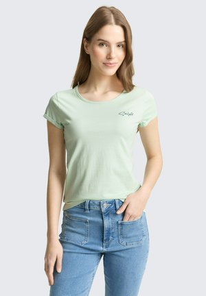 Młoda kobieta o jasnobrązowych włosach, ubrana w jasnozielony T-shirt z napisem "Smiles" oraz niebieskie dżinsy z wysokim stanem, pozująca z jedną ręką w kieszeni.