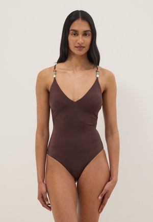 Femme aux longs cheveux foncés portant un maillot de bain une pièce marron avec des bretelles ornées de perles, debout devant un fond clair uni.