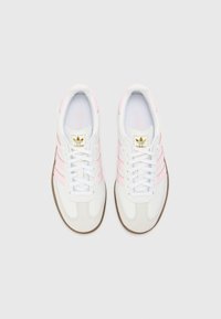 Sapatilhas brancas com riscas rosa e bico em camurça bege. Apresentam um logo dourado na língua e atacadores brancos. Sola de borracha texturizada.