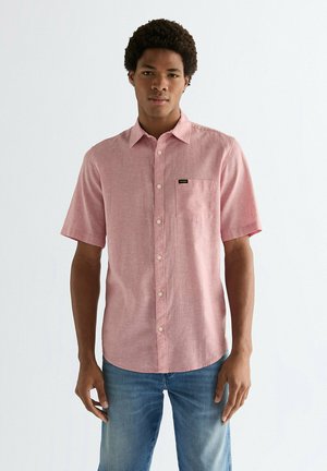 Jonge man draagt een roze overhemd met korte mouwen, een borstzak en een knoopsluiting, gecombineerd met een blauwe spijkerbroek, staand tegen een effen lichte achtergrond.