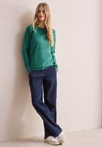 Maglione verde a maniche lunghe con una texture liscia, abbinato a jeans scuri e larghi e sneakers bianche. In piedi contro un muro chiaro.