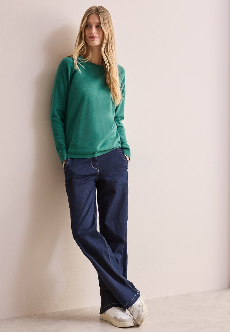 Maglione verde a maniche lunghe con una texture liscia, abbinato a jeans scuri e larghi e sneakers bianche. In piedi contro un muro chiaro.