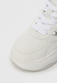 Bijela tenisica s teksturiranim mrežastim gornjim dijelom i glatkim sintetičkim kožnim detaljima, s okruglim pertlama i blago zakrivljenim gumenim potplatom.