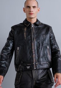Giacca in pelle nera con cerniera frontale, colletto e due tasche con cerniera. Texture consumata con leggere pieghe e un dettaglio quadrato sull'orlo.