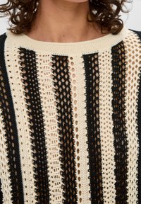 Zbliżenie osoby noszącej dziergany sweter z pionowymi czarnymi i kremowymi ażurowymi paskami.