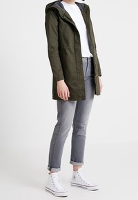 Chaqueta ligera de color verde oliva con capucha gris, cierre de cremallera y bolsillos laterales. Combinada con jeans gris enrollados y zapatillas altas blancas.
