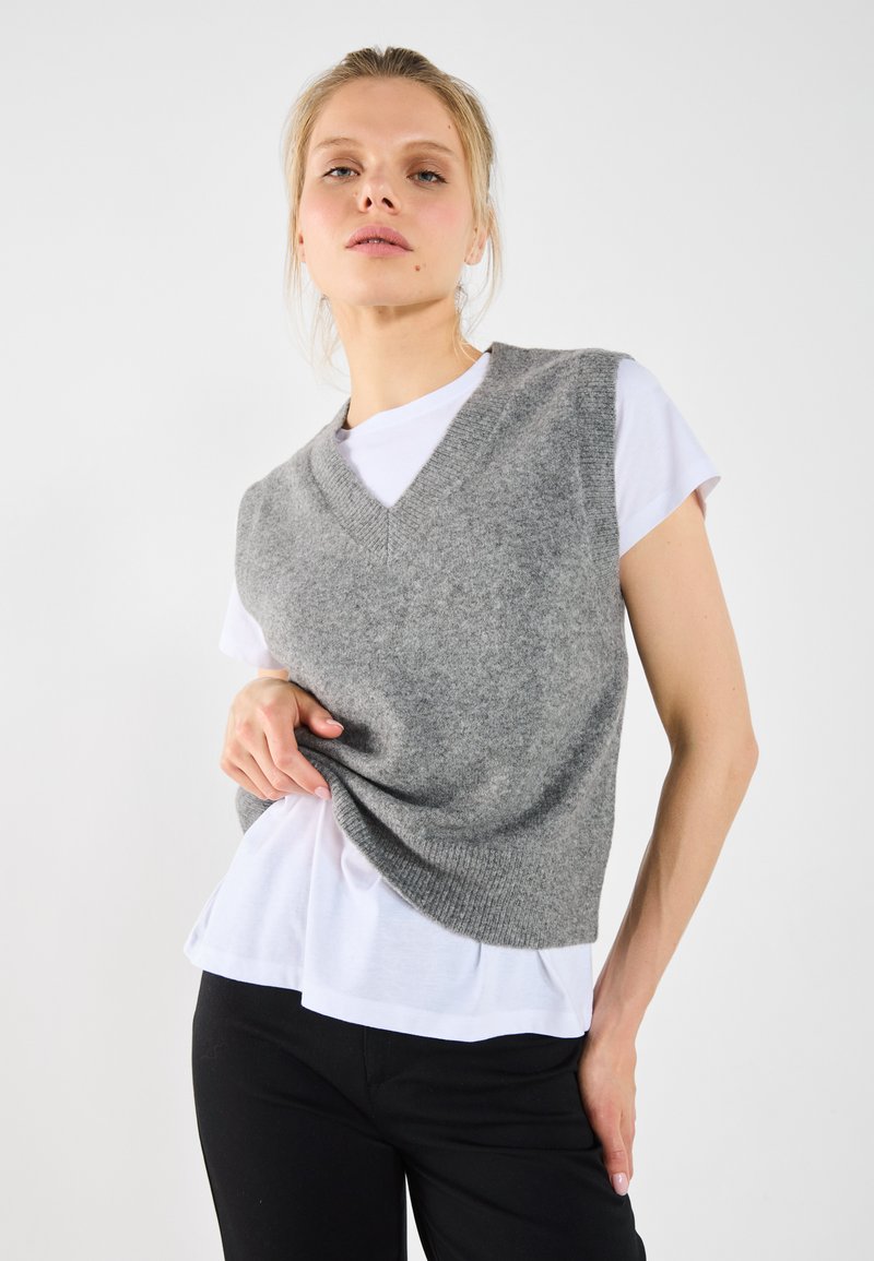 Grå ulltröja med V-ringning, lager på lager över en vit t-shirt. Snygg fit med ribbad kant, tillsammans med svarta byxor.