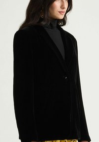 Blazer en velours noir avec un bouton unique, revers crantés et épaules structurées, présentant une texture lisse et une coupe ajustée.