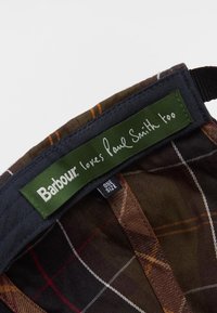 Καφέ και πράσινο καρώ καπέλο με πράσινη ετικέτα που λέει "Η Barbour αγαπά επίσης τον Paul Smith" σε λευκή γραφή. Περιλαμβάνεται ετικέτα ενός μεγέθους.