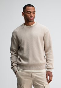 Beige gerippter Strickpullover mit Rundhalsausschnitt und langen Ärmeln. Strukturiert, lockere Passform, kombiniert mit hellbeigen Hosen.