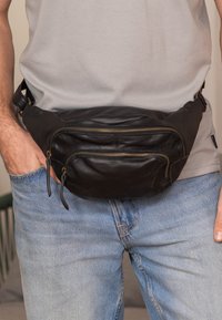 Schwarze Leder-Hüfttasche mit zwei Reißverschlüssen vorne und Lederziehstäbchen, getragen über einem grauen Shirt und blauen Jeans. Weiche Textur und geschwungene Form.