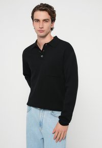 Czarna koszulka polo z długim rękawem wykonana z miękkiego materiału, z kołnierzykiem zapinanym na guziki, kieszenią na piersi, zestawiona z jasnoniebieskimi dżinsami.