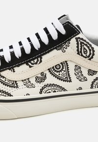 Canvas sneakers har en kremfarget base, svarte paisleymønstre, hvite snøringer og en svart semsket trim. Gummisåle med svart aksent.