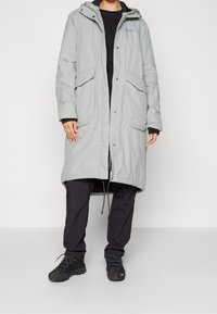 Veste longue gris clair avec capuche, dotée de deux poches avant, de boutons-pression et d'un cordon de serrage à l'ourlet. Portée sur des vêtements noirs.