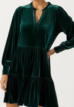 Robe en velours vert foncé à manches longues avec volants froncés, encolure en V et poignets avec revers portée par une personne avec une main sur la hanche.