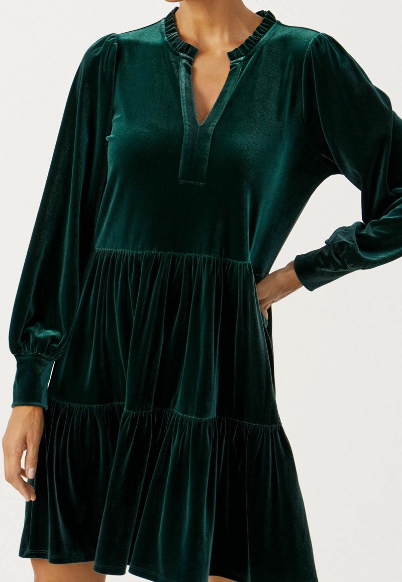 Robe en velours vert foncé à manches longues avec volants froncés, encolure en V et poignets avec revers portée par une personne avec une main sur la hanche.