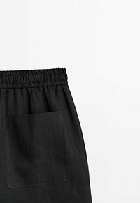 Pantalon en tissu noir avec taille élastique et grande poche carrée sur le côté droit sur fond blanc.