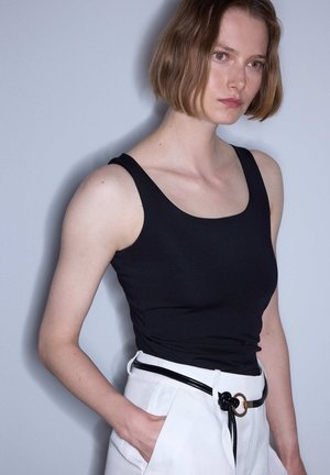 Donna con capelli corti castani che indossa un top nero senza maniche e pantaloni bianchi con cintura nera, in piedi davanti a uno sfondo grigio chiaro.