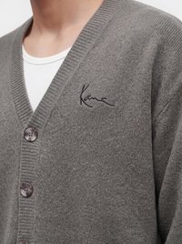 Close-up af en grå strikket cardigan med tre knapper, båret over en hvid skjorte, med en lille broderet sort signatur på venstre bryst.