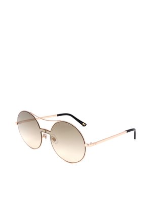 Occhiali da sole oversize tondi con sottile montatura in metallo oro, lenti sfumate marroni e punte delle aste nere, inclinati verso destra su sfondo bianco.