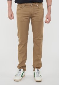 Chinos beige in cotone con un taglio slim, design a cinque tasche, chiusura frontale con bottone e una texture leggermente sbiadita. Abbinati a sneakers bianche e verdi.