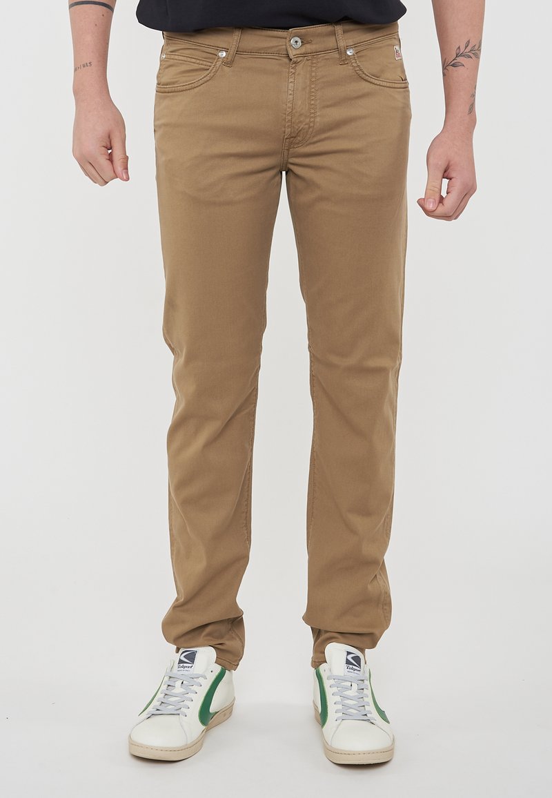 Chinos beige in cotone con un taglio slim, design a cinque tasche, chiusura frontale con bottone e una texture leggermente sbiadita. Abbinati a sneakers bianche e verdi.