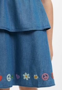 Jeanskleid mit schichtweisem Design, verziert mit farbenfrohen gestickten Mustern, darunter Blumen und Friedenssymbole am Saum.