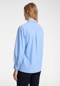 Camicia colletto azzurro chiaro con strisce verticali bianche, maniche lunghe e polsini con bottone. Presenta un dettaglio con piega sul retro per una maggiore libertà di movimento.