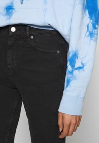 Mustat farkut, joissa on istuva malli, hopeinen nappi ja taskuyksityiskohdat, yhdistettynä vaaleansinaiseen tie-dye huppariin.