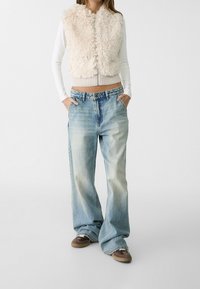 Mujer vistiendo un chaleco corto de peluche beige sobre una camiseta de manga larga blanca, jeans claros holgados y zapatillas marrones, de pie con las manos en los bolsillos.