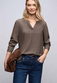 Brun langærmet bluse med V-hals design, parret med blå denimjeans. En brun ruskinds clutchnik holdes i den ene hånd.