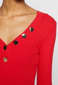 Haut en maille côtelée rouge avec un décolleté en V profond, agrémenté de boutons métalliques le long de l'encolure et de manches longues. La texture est douce et lisse.