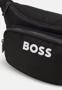 Crna platnena torba s remenom koja ima dva džepa na zatvarač, teksturiranu površinu i 'BOSS' logo u podebljanim bijelim slovima sprijeda.