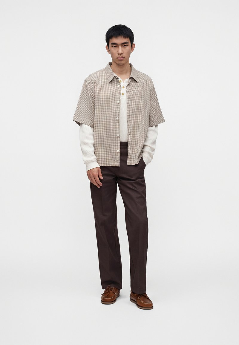 Giovane uomo in piedi, indossa una camicia beige a quadri a maniche corte sopra una maglia bianca a maniche lunghe, pantaloni marrone scuro e scarpe di pelle marrone.