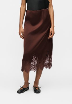 Donna che indossa una gonna midi di raso marrone scuro con orlo in pizzo, abbinata a ballerine nere, in piedi davanti a uno sfondo semplice.