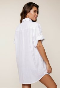 Abito camicia bianco con maniche arrotolate, caratterizzato da una sottile trama a righe e un orlo curvo, progettato per una vestibilità ampia.