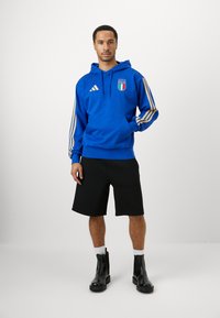 Μπλε φούτερ με χρυσές ρίγες και σήμα Ιταλίας, συνδυασμένο με μαύρες κοντές παντελόνες και μαύρες μποτίνες. Οι κάλτσες είναι λευκές, τα υποδήματα έχουν γυαλιστερή φινίρισμα.