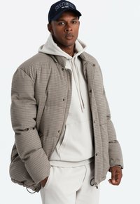 Beige en bruine geruite gewatteerde jas, gedragen over een witte hoodie. Voorzien van drukknoopsluitingen en elastische manchetten. Donker pet op het hoofd.