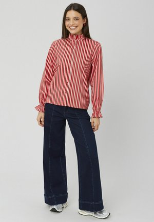 Chemise à rayures rouges et blanches avec col haut, manches longues et poignets volantés ; associée à un pantalon large en denim foncé et des baskets.