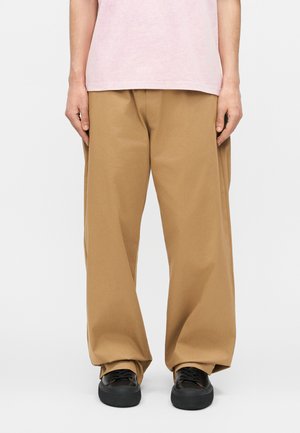 Άτομο που φοράει φαρδιά παντελόνια σε καμηλό χρώμα, μαύρα αθλητικά παπούτσια και ανοιχτό ροζ πουκάμισο, στέκεται μπροστά σε λευκό φόντο.