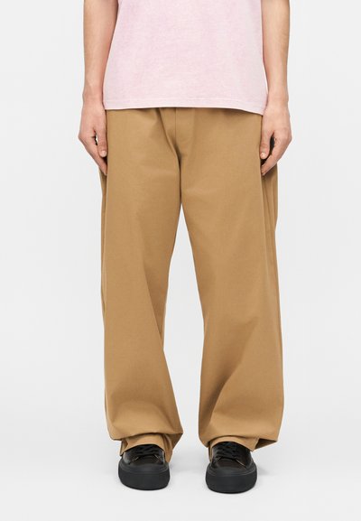 Personne portant un pantalon ample de couleur camel, des baskets noires et une chemise rose clair, debout devant un fond blanc.