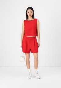 Haut court sans manches rouge associé à un short taille haute rouge. Le mannequin porte un petit sac blanc et des baskets montantes blanches.